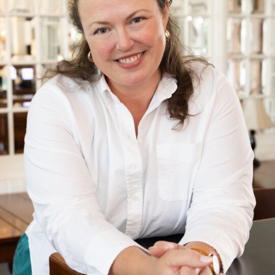 Headshot of Sarah Staiger. Leaning forward on a counter with hands clasped in front.